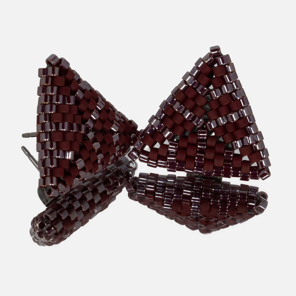 Pair of small hand-woven beaded triangle earrings in matte and glossy burgundy, standing on a mirror