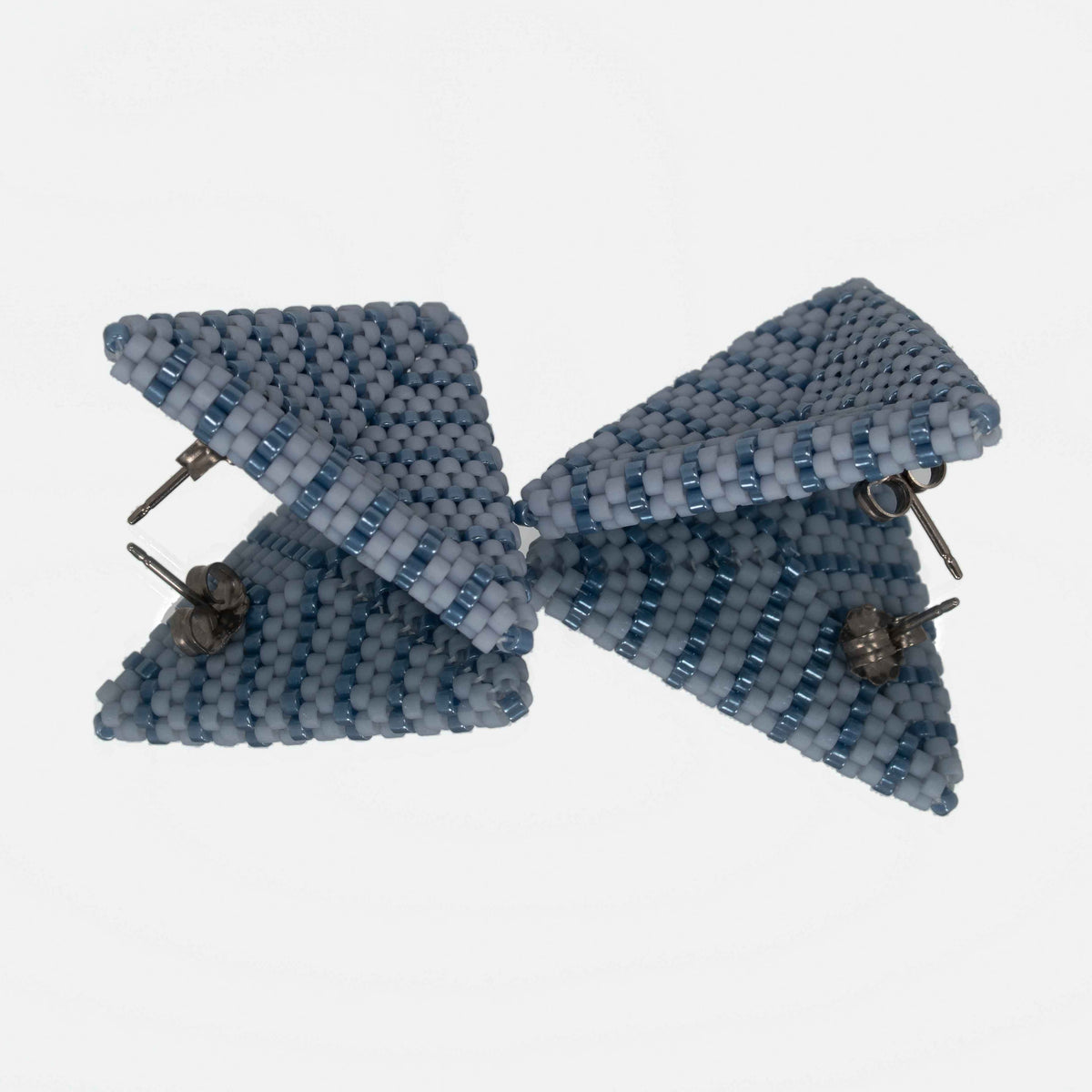 Pair of dusty blue and denim beaded triangle earrings facing each other on a mirror, showing the titanium posts and the woven edges