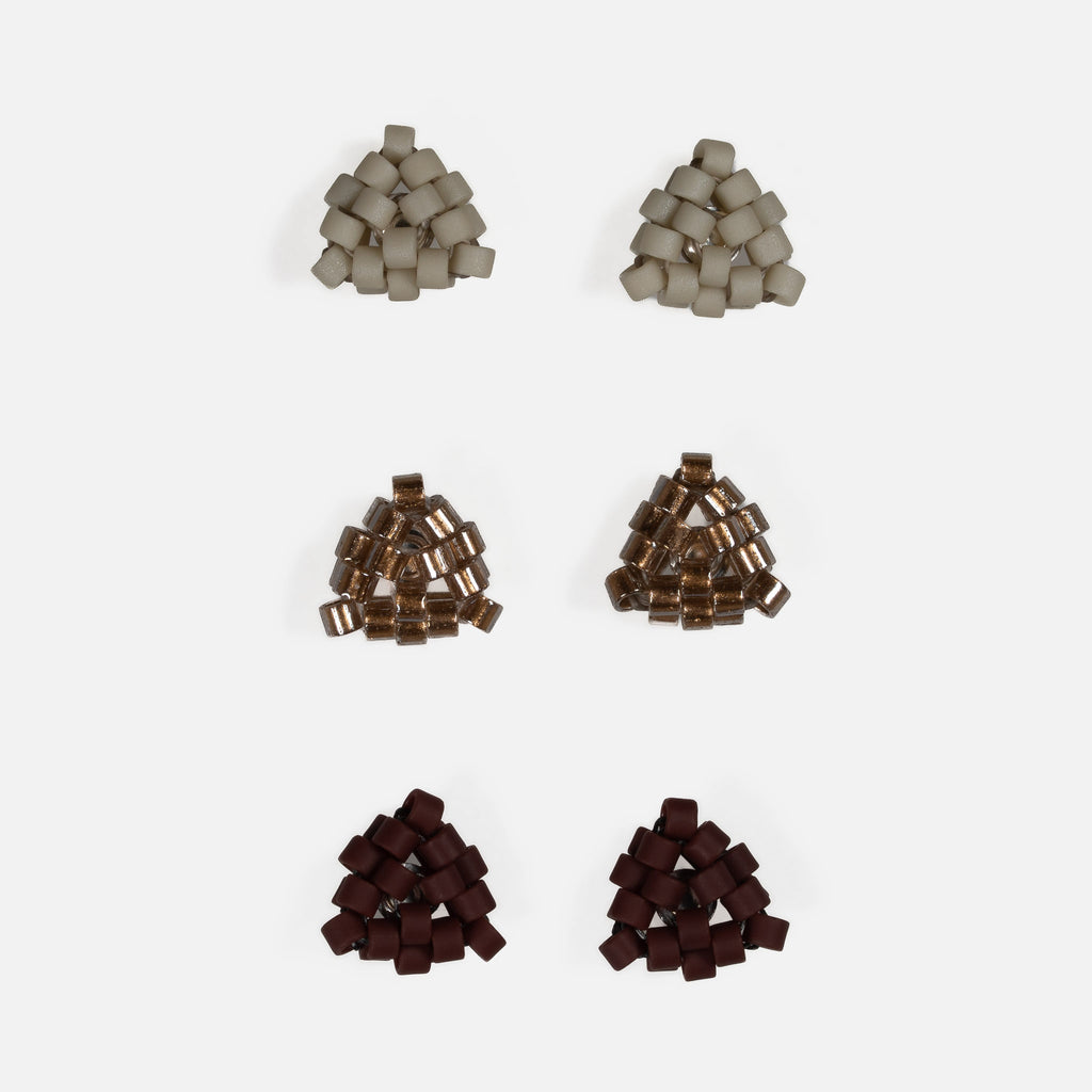 Three pairs of tiny triangular beaded stud earrings in matte greige, antique copper-lined glass, and matte oxblood, arranged in rows on a light gray background